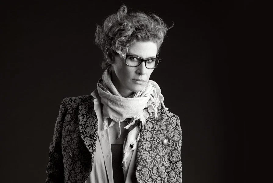 Schwarz-Weiß-Foto einer Frau mit kurzen, lockigen Haaren, trägt eine Brille, einen Schal und eine strukturierte Jacke, Blick in die Kamera