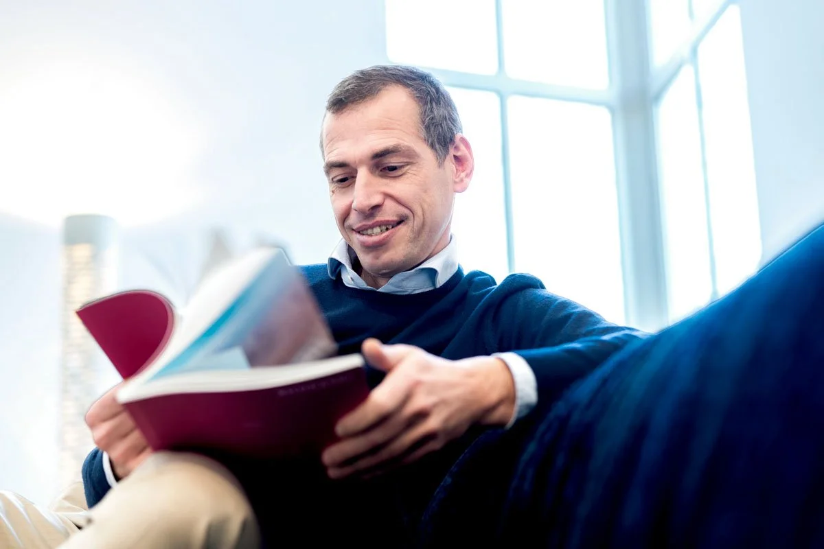 Ein Mann liest ein Buch in einem hell erleuchteten Raum mit großen Fenstern.
