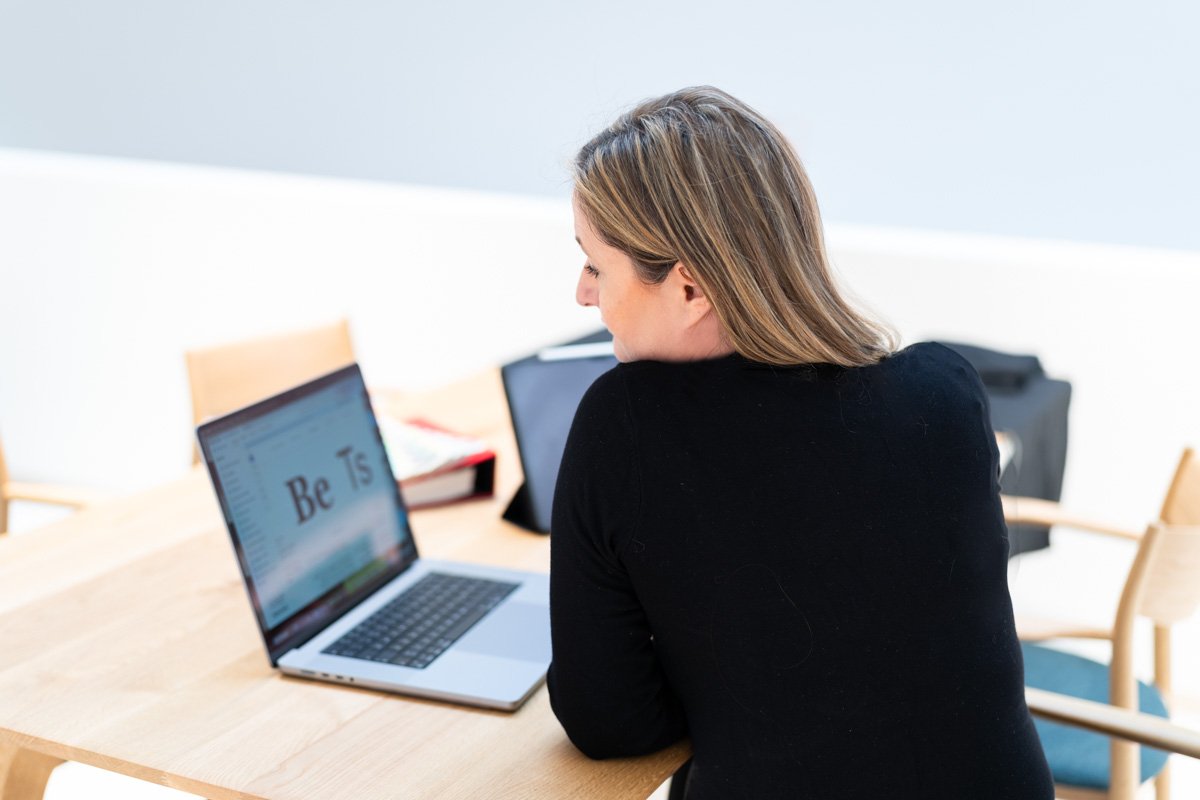 Eine Frau sitzt an einem Tisch und arbeitet an einem Laptop in einem hellen Raum.