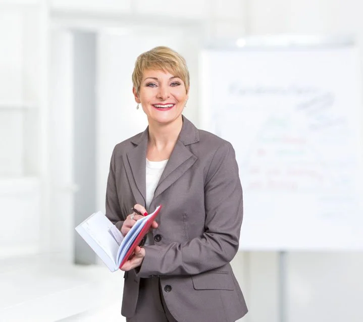 Lächelnde Frau in einem grauen Anzug hält einen Notizblock in der Hand, im Hintergrund eine weiße Tafel.