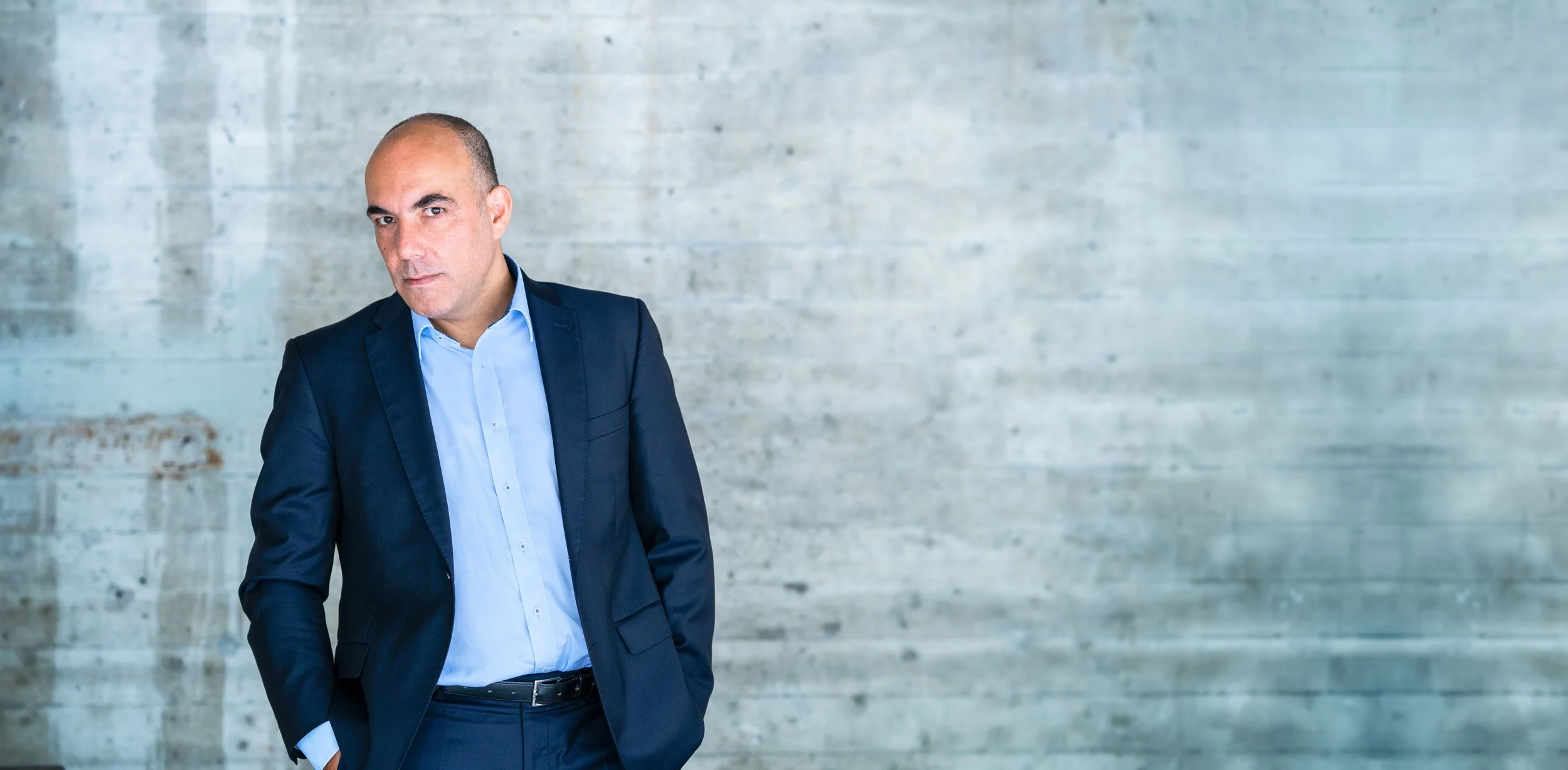 Ein Mann in dunklem Anzug steht vor einer leeren, grauen Betonwand.