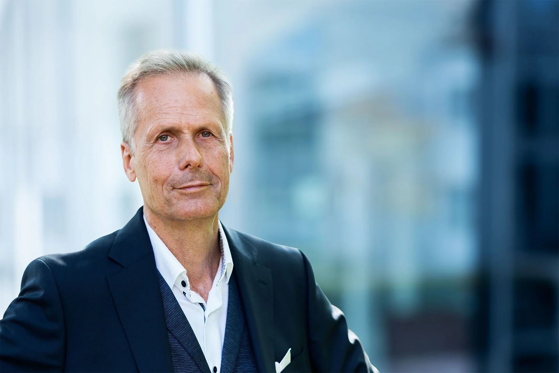 Ein älterer Mann in dunklem Anzug und weißem Hemd steht vor einer modernen Büroumgebung mit Glasfenstern.