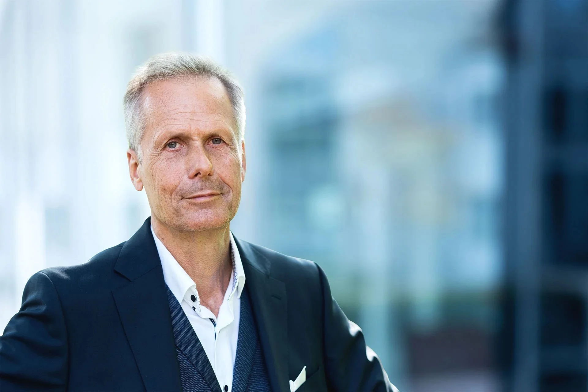 Ein älterer Mann in dunklem Anzug und weißem Hemd steht vor einer modernen Büroumgebung mit Glasfenstern.