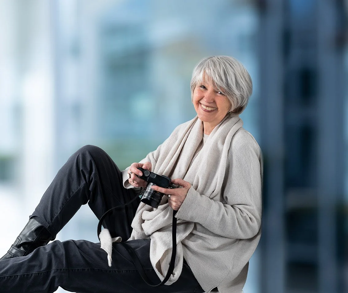 Ältere Frau mit kurzen grauen Haaren sitzt lachend, hält eine Kamera in den Händen, trägt einen hellen Pullover, ein helles Tuch und schwarze Hose, im Hintergrund ist eine unscharfe moderne Gebäudefassade.