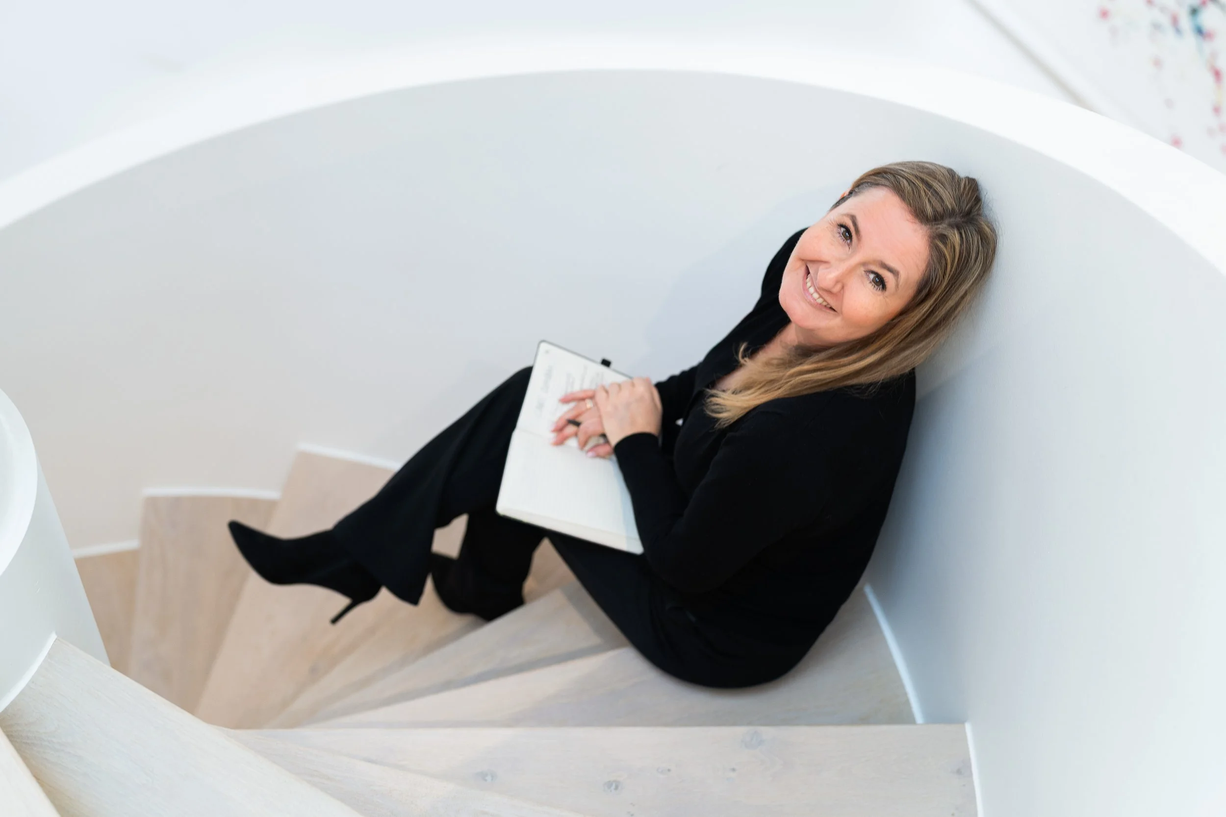 Eine lächelnde Frau in schwarz sitzt auf einer Treppe, sieht nach oben und hält eine Notizbuch in den Händen.