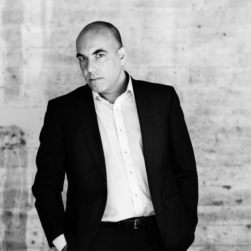 Schwarz-weiß-Foto eines Mannes in einem schwarzen Anzug und weißem Hemd, der nach links schaut, vor einer Wand aus Holzbohlen.