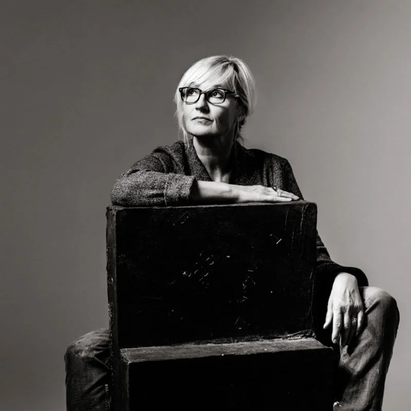 Schwarz-Weiß-Foto einer älteren Frau mit Brille, die auf einem Stuhl sitzt und nach oben schaut.