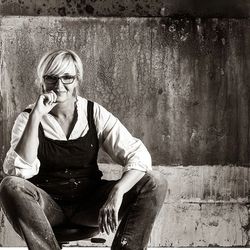 Frau sitzt entspannt vor einer alten, verfallenen Wand mit sichtbaren Rissen, trägt Brille, weiße Bluse und schwarze Schürze, lächelt leicht.