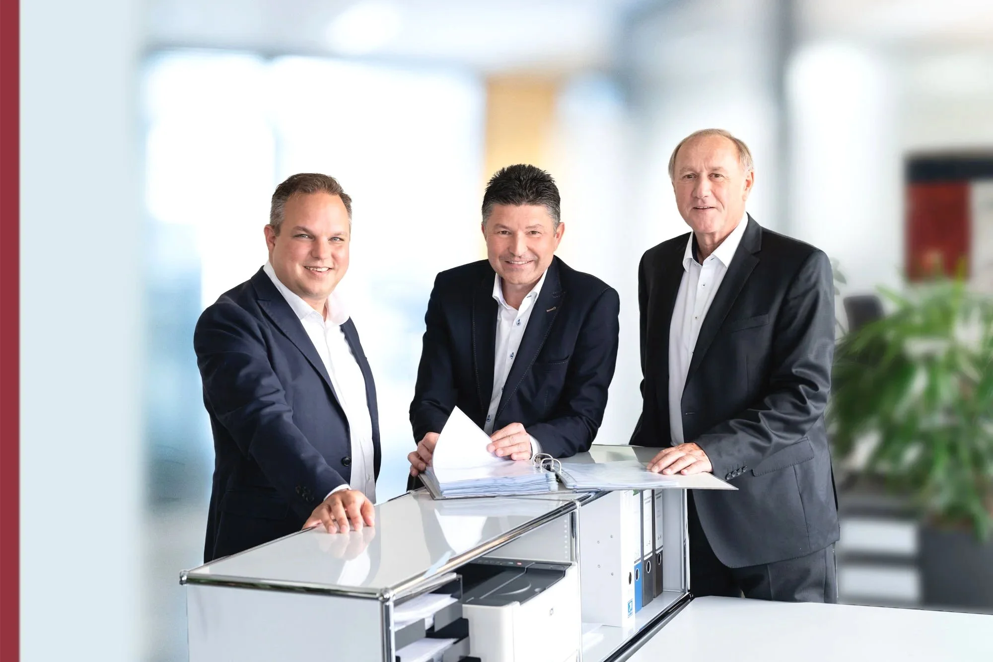 Drei Männer in Anzügen stehen um einen Schreibtisch mit Aktenordnern, in einem modernen Büro.
