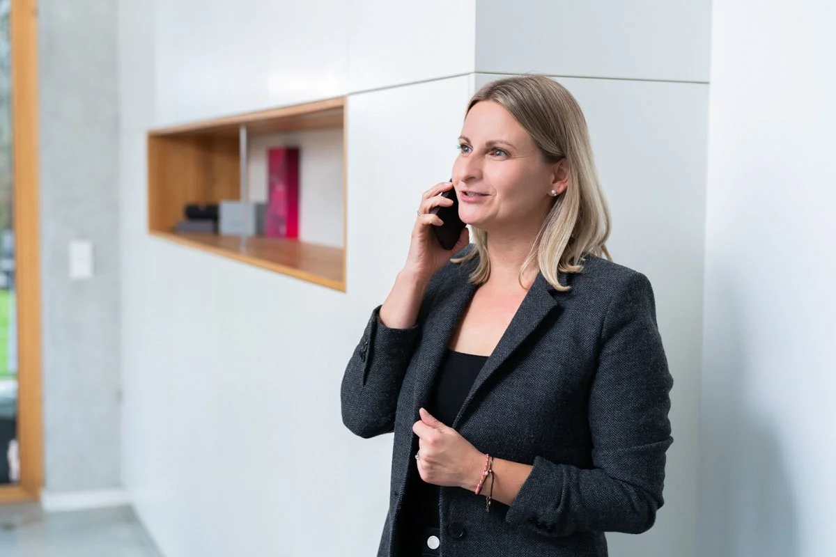 Eine Frau im schwarzen Blazer spricht am Telefon in einem modernen Büro.