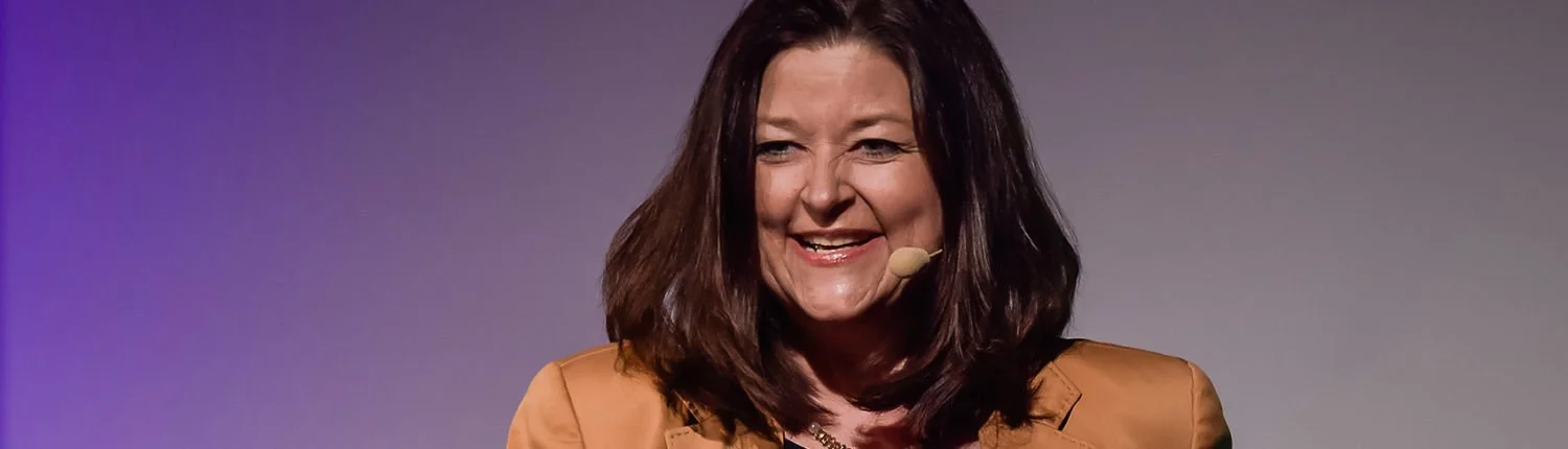 Eine lachende Frau mit braunem Haar, die ein Anzug trägt, vor einem grauen Hintergrund, spricht in ein Mikrofon.