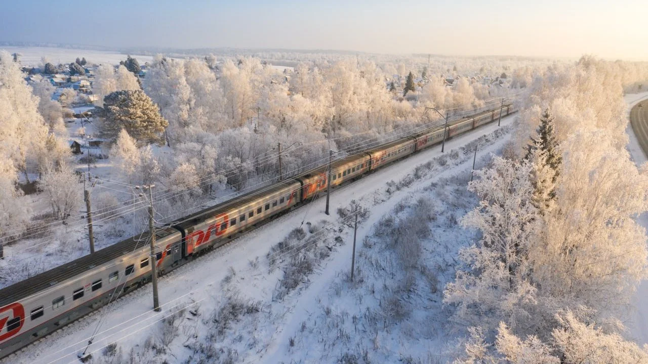 An aerial view of a snow-covered landscape with trees and houses, showing a long train traveling on an electric railway track.