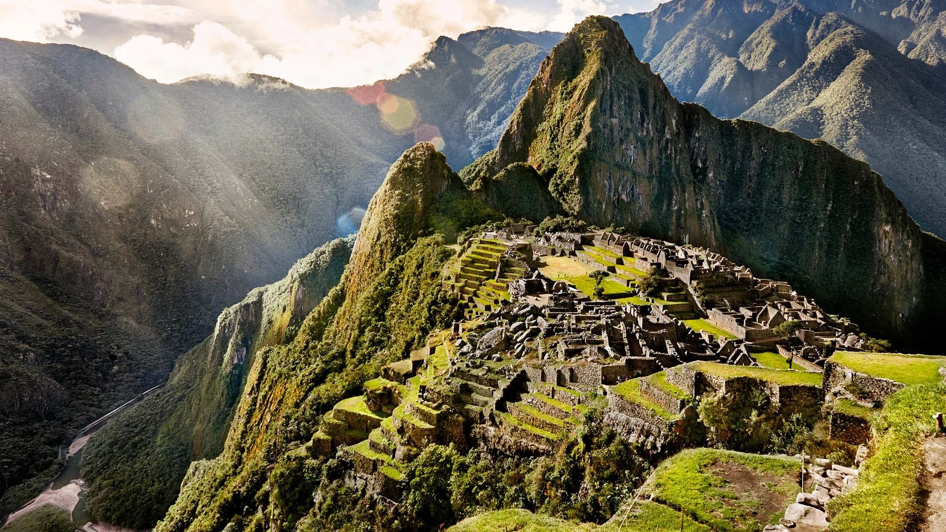 Ancient Incan city of Machu Picchu built on mountain ridge surrounded by lush green valleys and high peaks, with terraced stone structures and ceremonial sites under a partly cloudy sky.