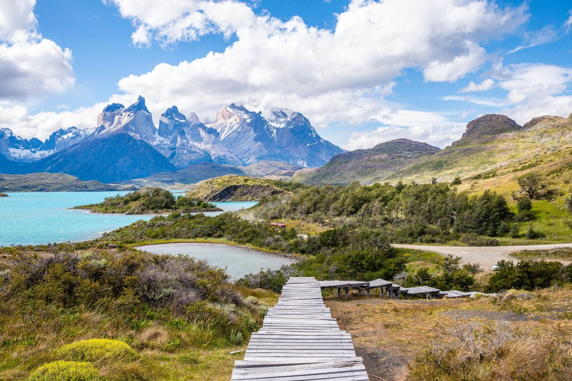 Wooden pathway leading through green grass and shrubs, with lakes and mountains with snow-capped peaks in the background under a partly cloudy sky.