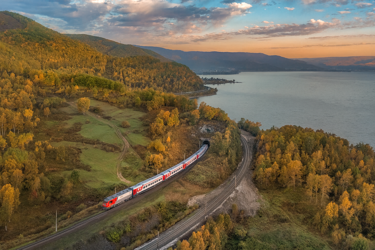 A passenger train travels through a scenic landscape with autumn foliage along a river and hills at sunset.