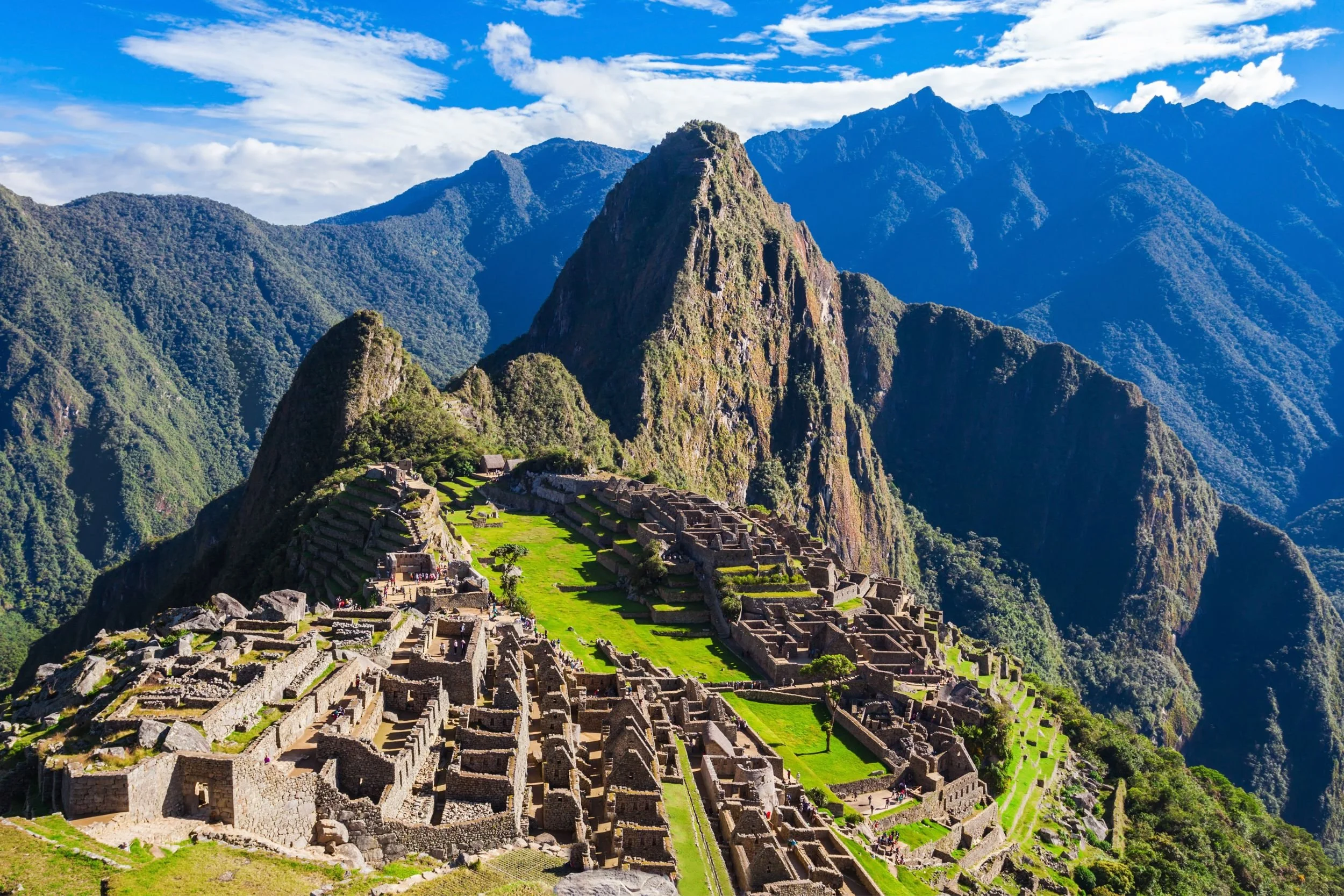 Ancient Incan ruins on a mountain with a prominent peak, surrounded by lush green and misty mountains in the background, under a partly cloudy sky.