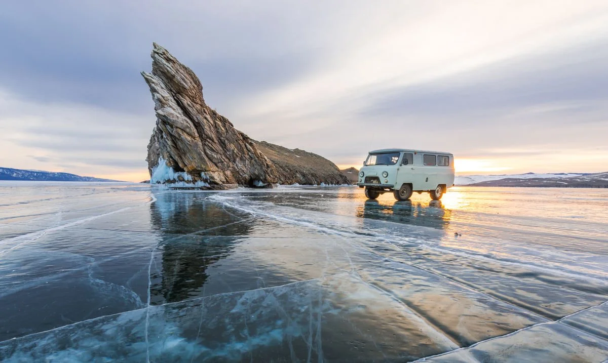 A vintage van parked on a frozen lake with a large rock formation in the background during sunset.