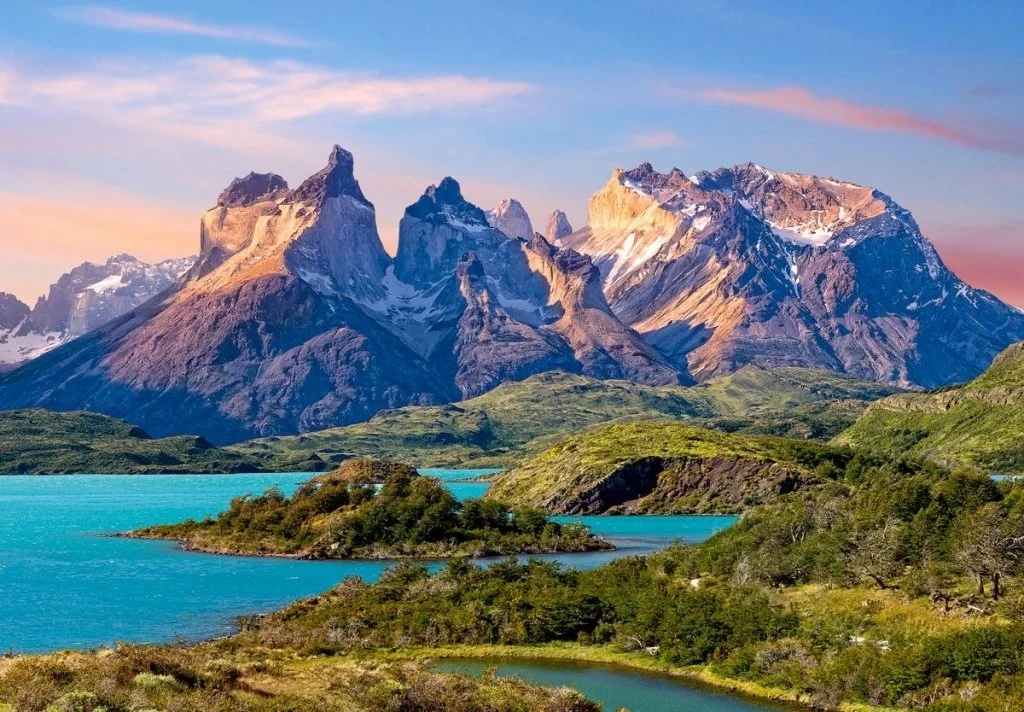 Scenic landscape of rugged mountains with snow patches, green hills, and turquoise lakes during sunset or sunrise.