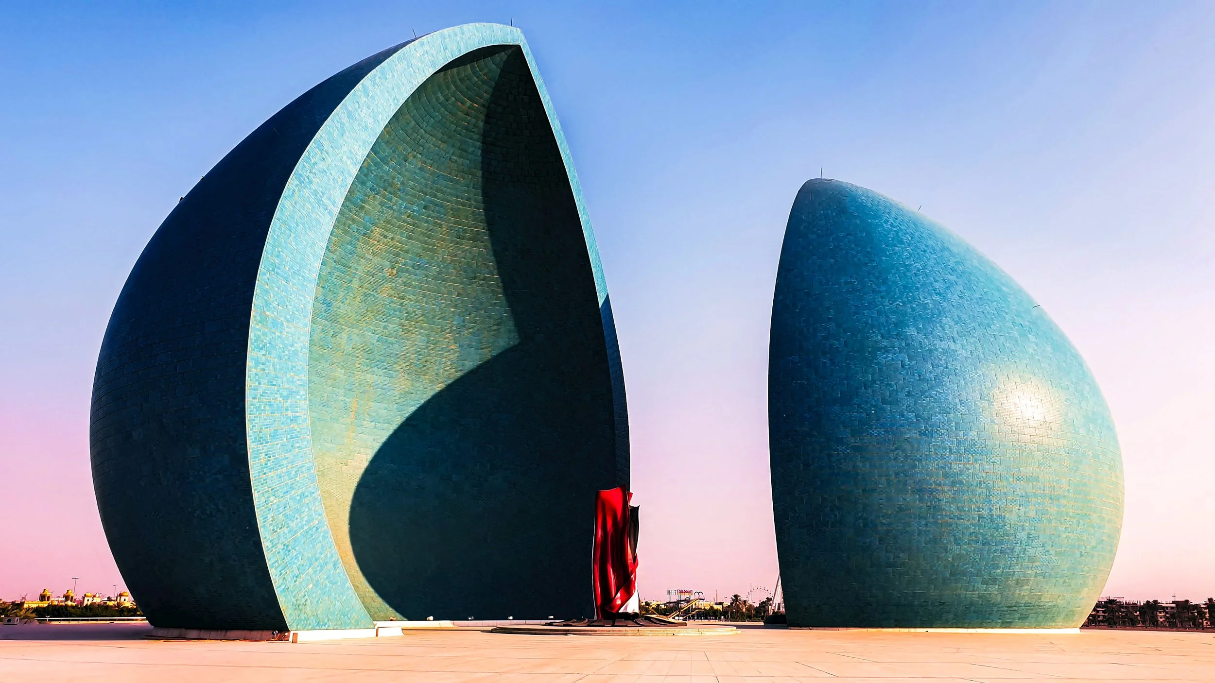 Modern architectural building with two large, curved, blue-tiled structures resembling shells, connected by a red and black sculpture in front, under a clear blue sky.