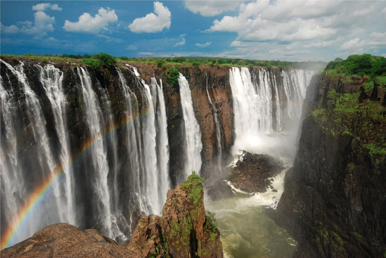 Multiple waterfalls cascading down a rocky cliff with green vegetation, a rainbow visible in the mist, and a cloudy sky above.