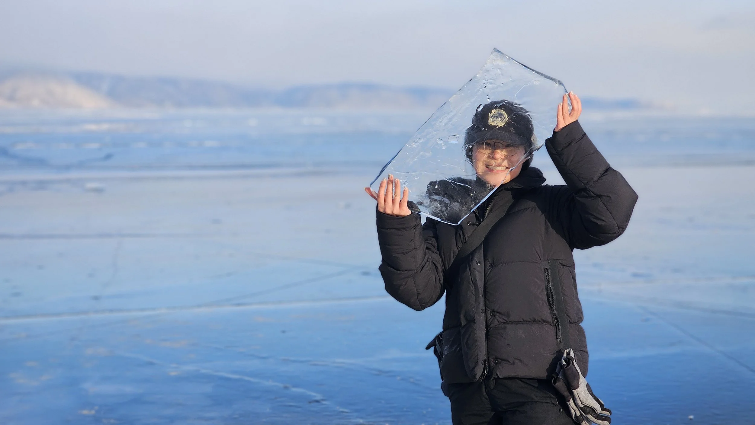 Person dressed in black winter jacket, hat, and glasses holding a piece of ice in an icy landscape with snow and distant mountains.