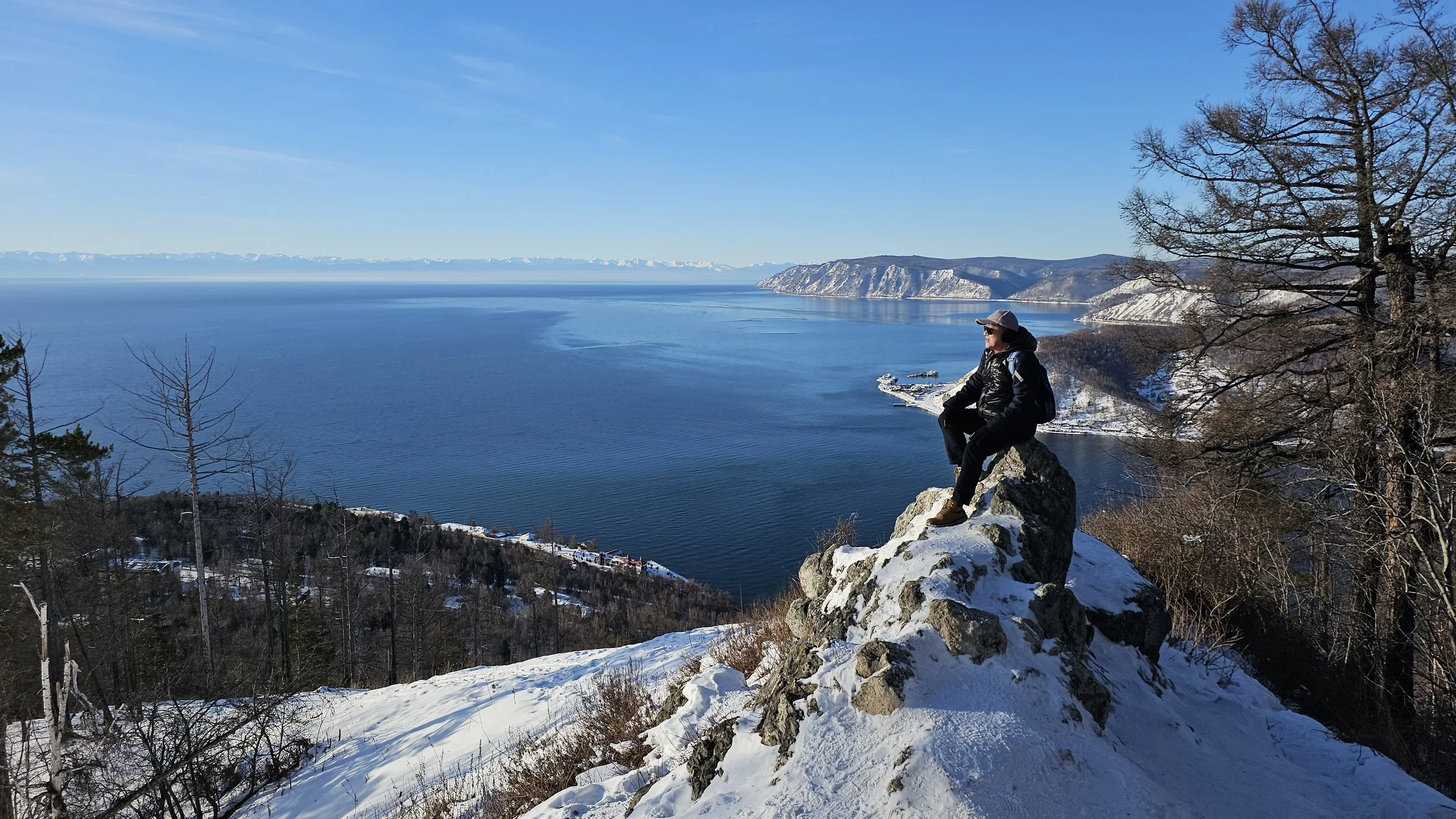 A person sitting on a rocky outcrop overlooking a snowy landscape and a large body of water under a clear blue sky.