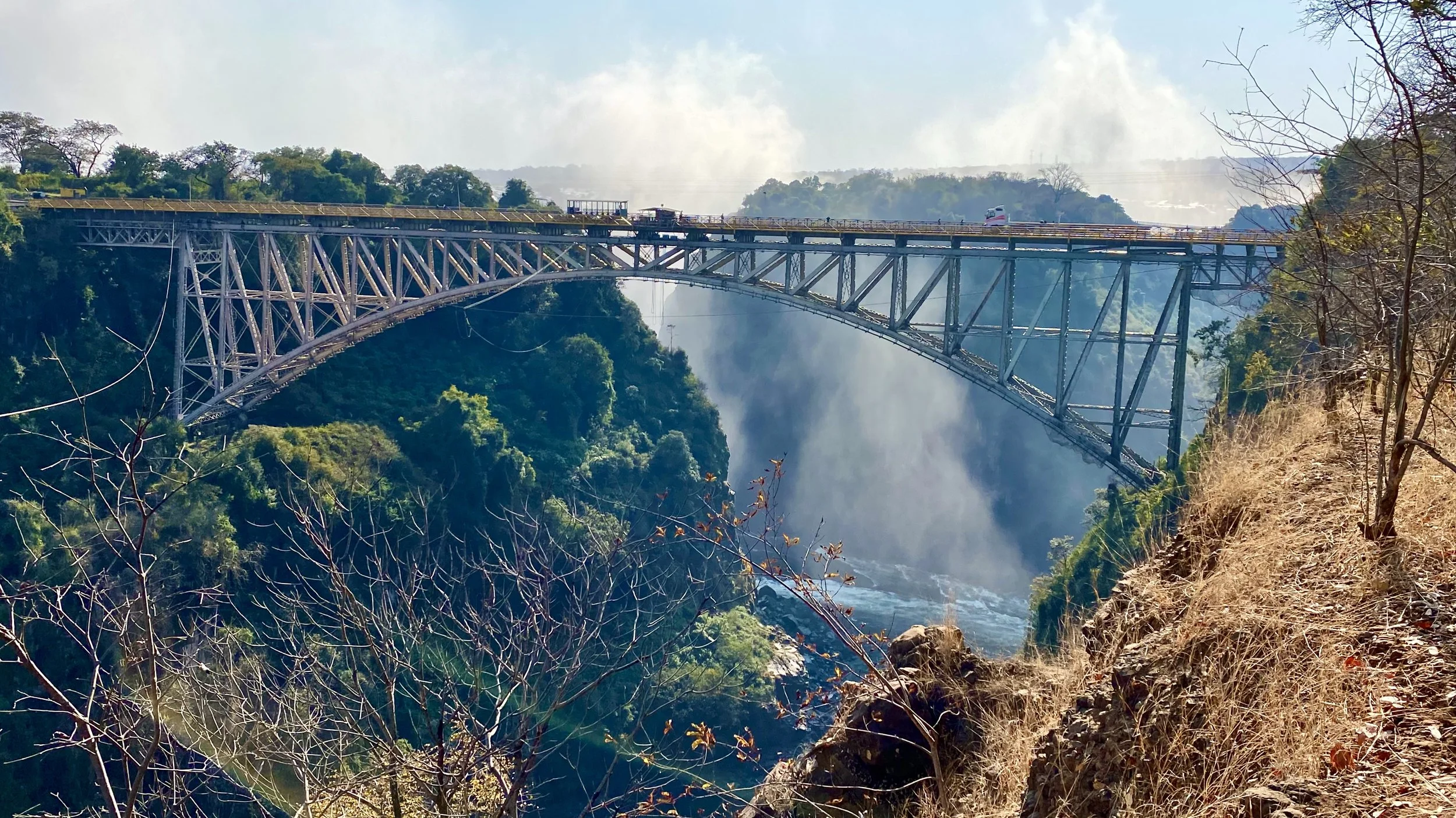 Steel arch bridge over a large waterfall in a lush green canyon with mist and trees.