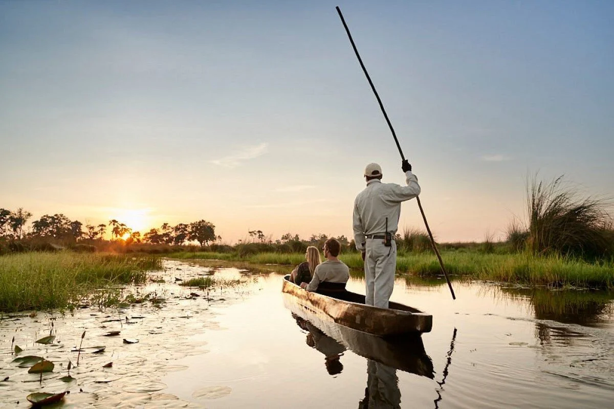 A boat with three people on a waterway at sunset, with a boat guide standing and holding a pole, while two passengers sit and look around amid green plants.