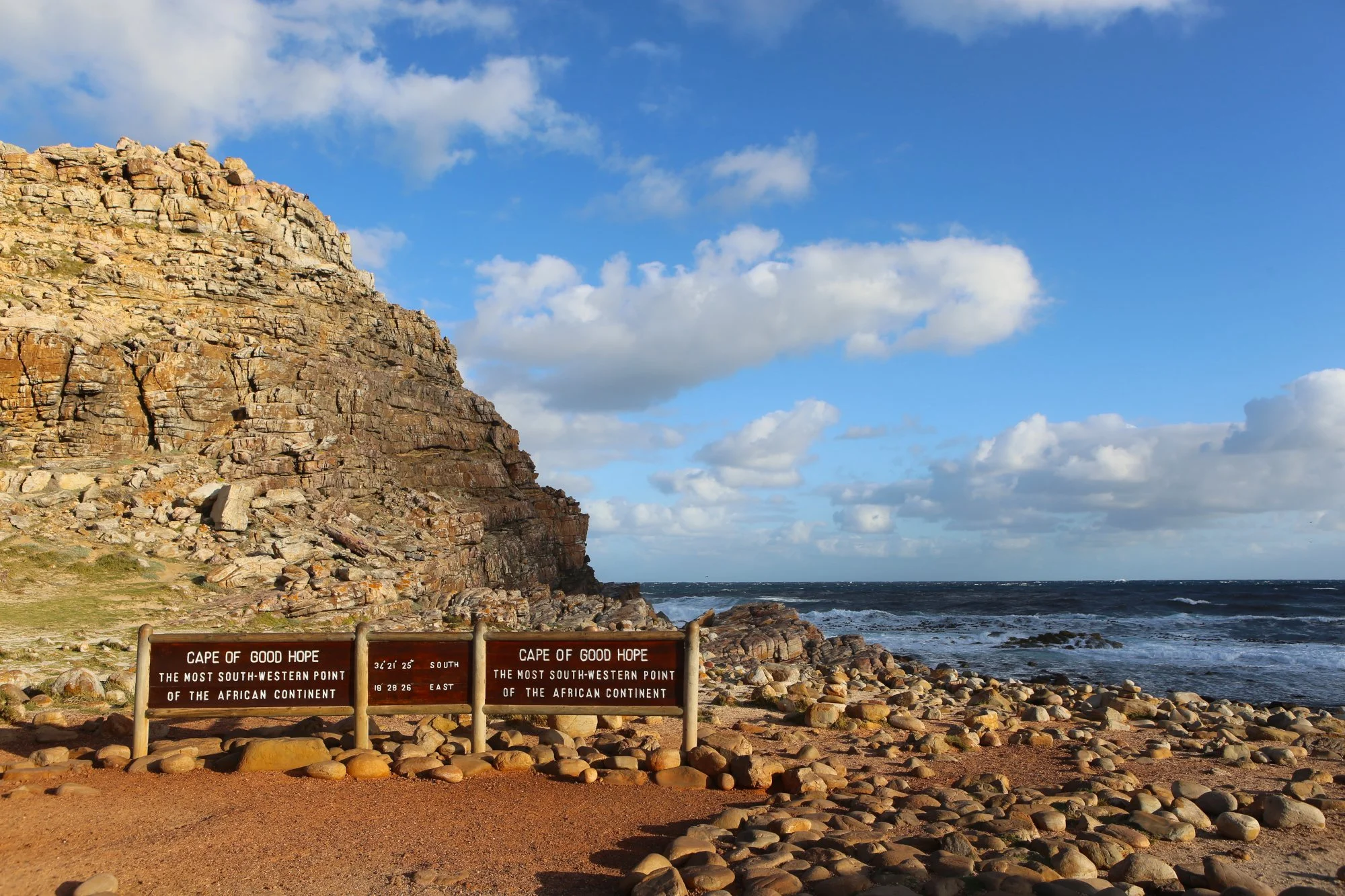 View of the Cape of Good Hope, with rocky cliffs on the left, a rocky beach in the foreground, and ocean waves under a partly cloudy sky. There are signs indicating it is the Cape of Good Hope, the most south-western point of the African continent.