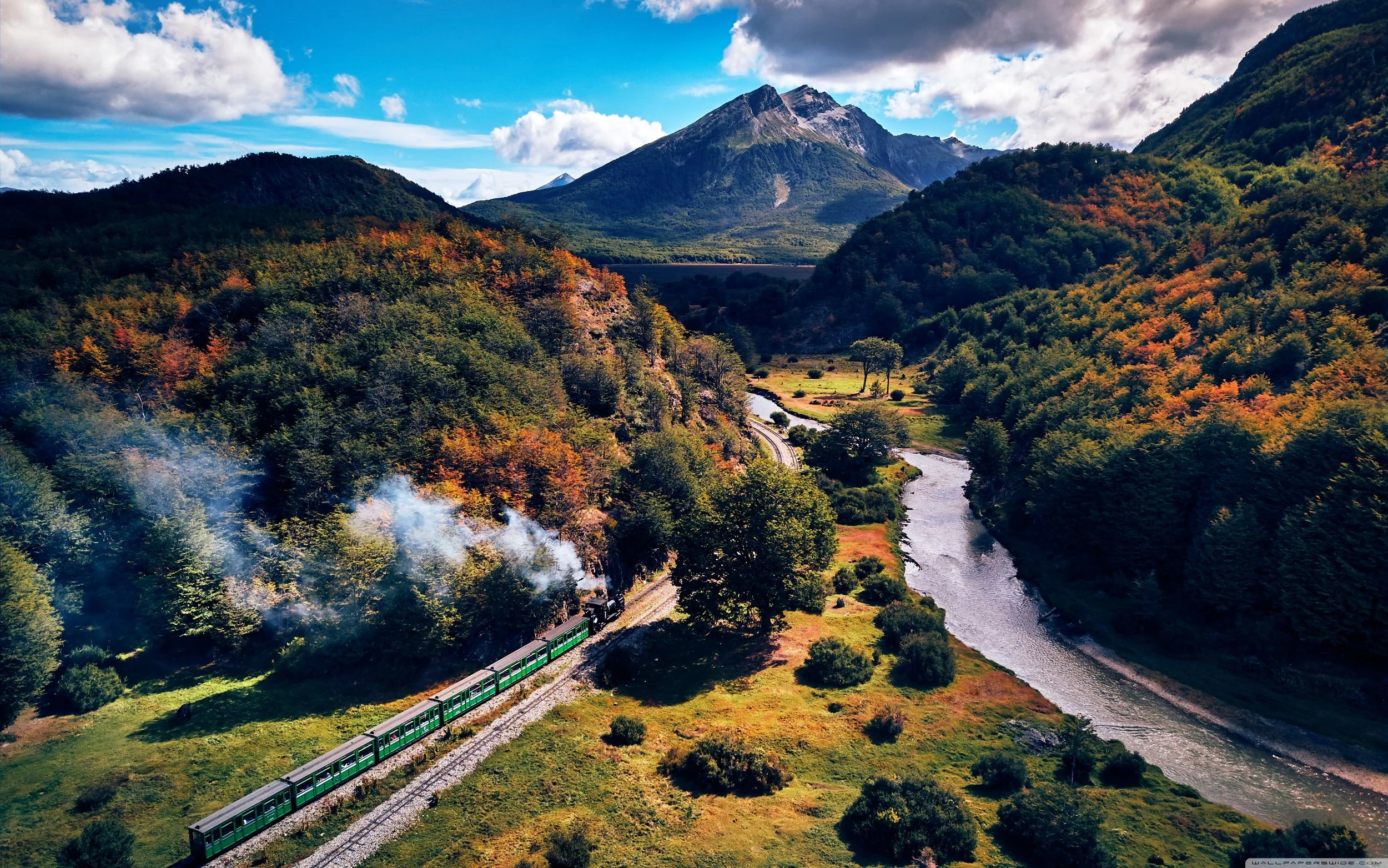 A train traveling through a lush, mountainous landscape with a winding river, colorful autumn trees, and a mountain in the background under a partly cloudy sky.