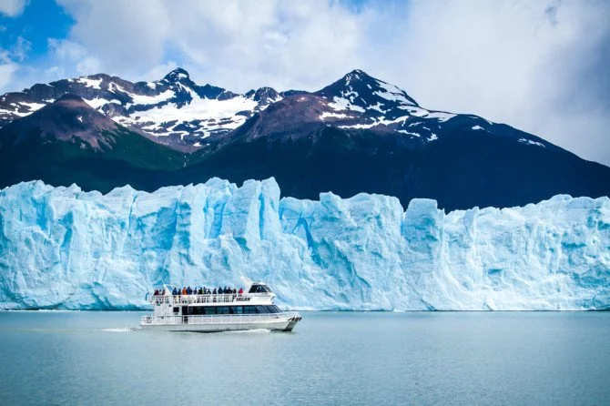 A large glacier with tall ice cliffs behind a boat on calm water with snow-capped mountains in the background.
