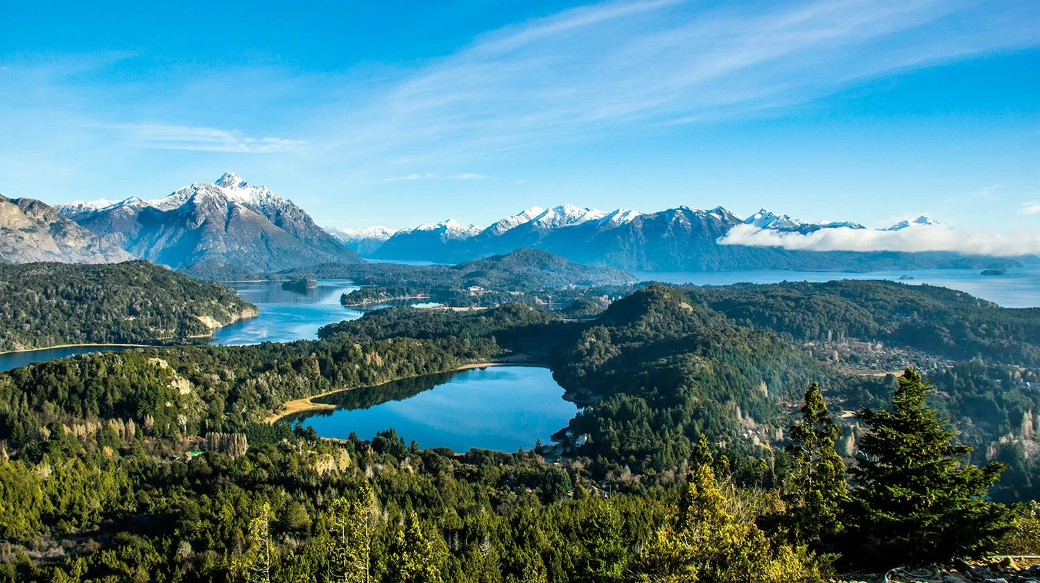 Scenic view of a landscape with multiple lakes surrounded by green forests, with snow-capped mountains in the background under a partly cloudy sky.