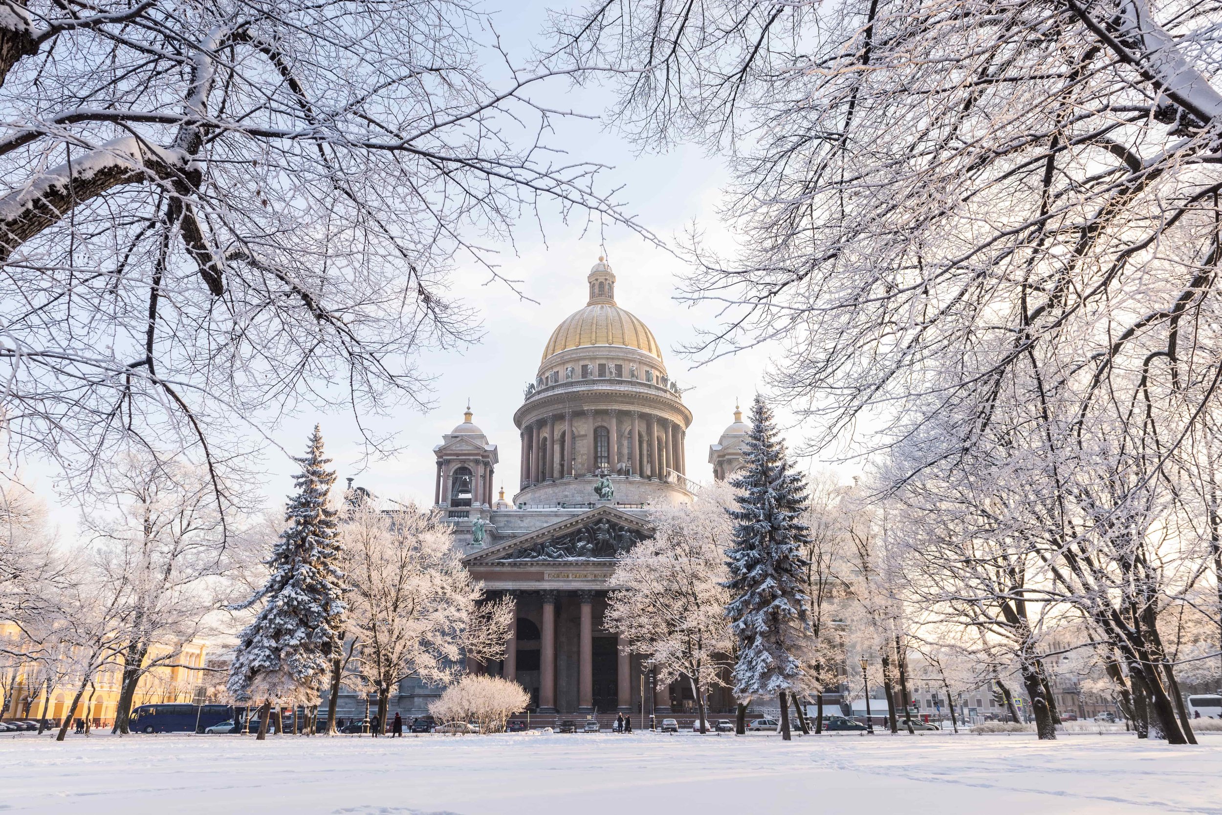 A grand historic building with a large dome surrounded by snow-covered trees and a snow-covered park area in the foreground.