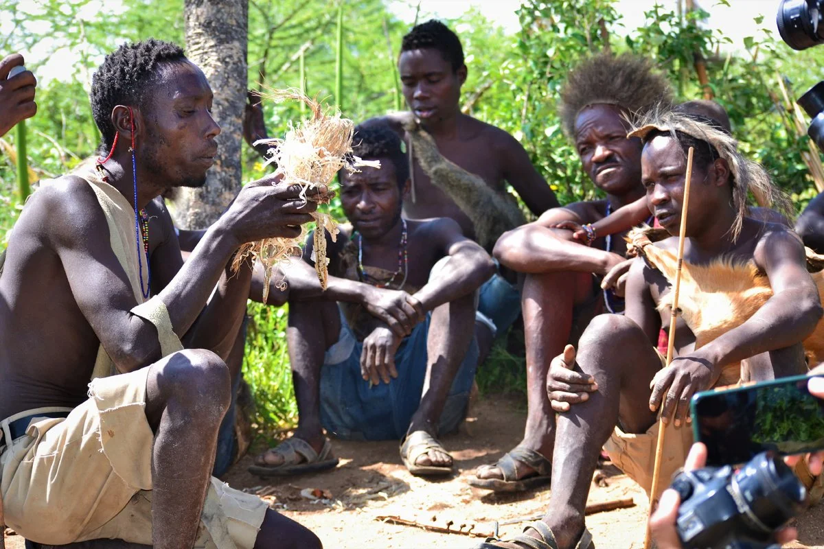 Group of indigenous people sitting on the ground outdoors in a forest, with some holding traditional tools and wearing minimal clothing, engaging in a cultural activity.