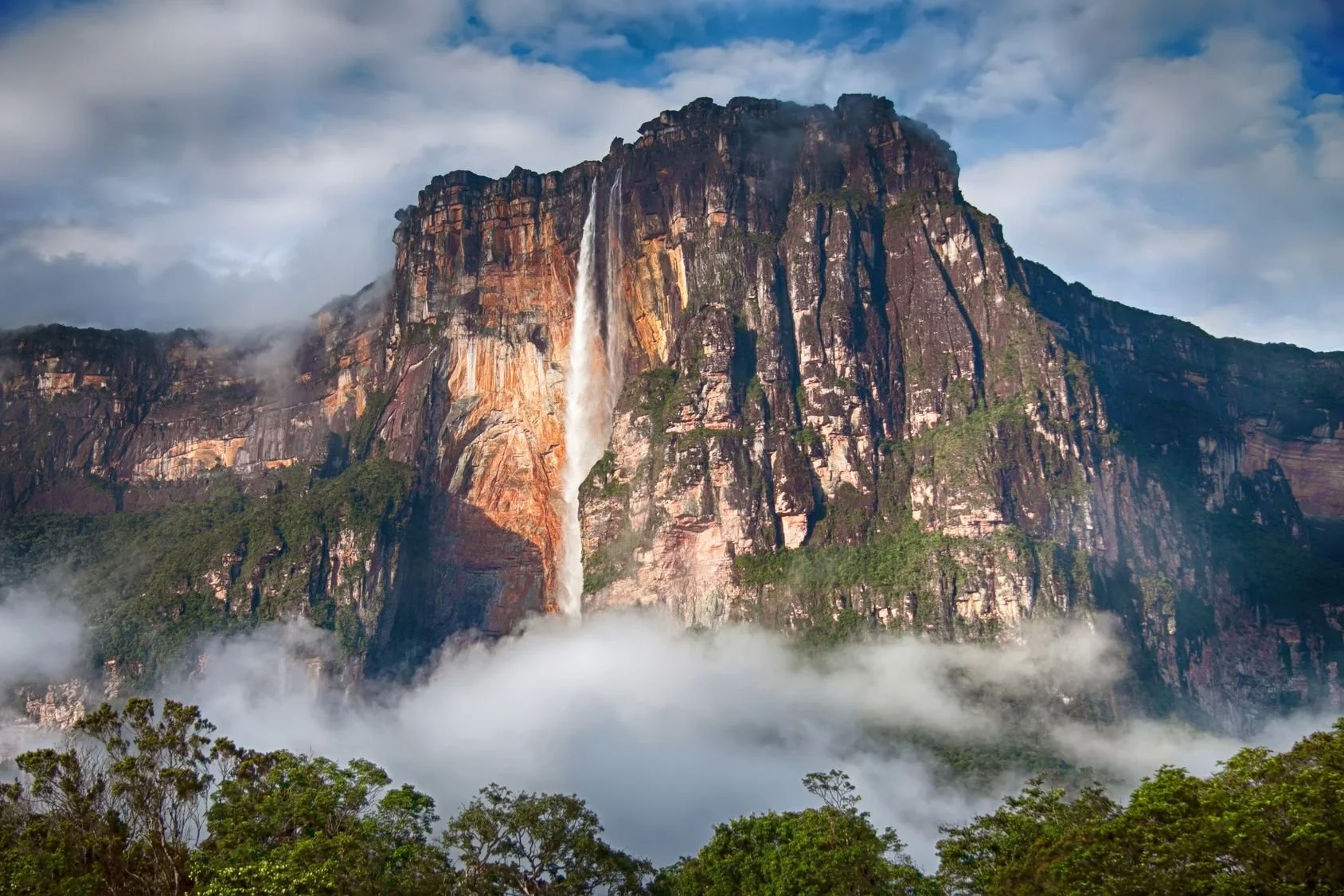 A large mountain with a waterfall flowing down its rocky surface, surrounded by clouds and green trees at the base.