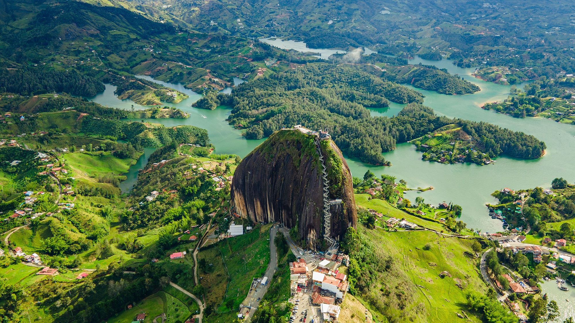 Aerial view of Sugarloaf Mountain overlooking a lush green landscape with small houses, winding roads, and a large body of water surrounding the mountain.