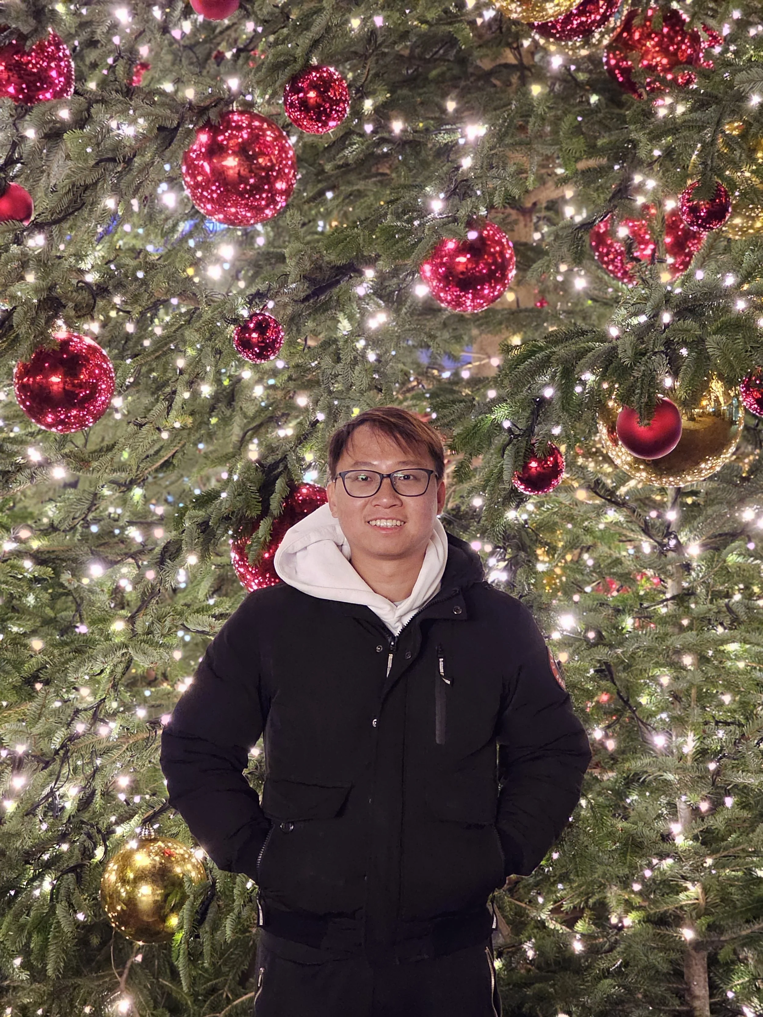 A person smiling and wearing glasses, a white hoodie, and a black jacket, standing in front of a decorated Christmas tree with red and gold ornaments and white lights.