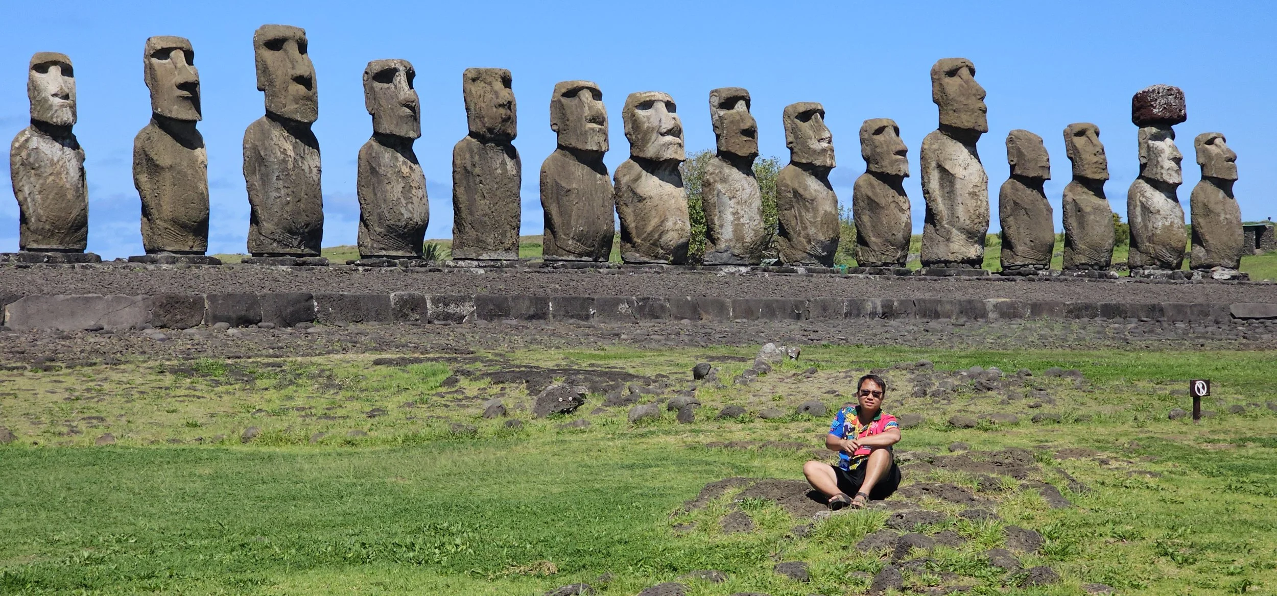 A person sitting on the grass in front of the Moai statues at Easter Island, with the statues towering behind against a bright blue sky.