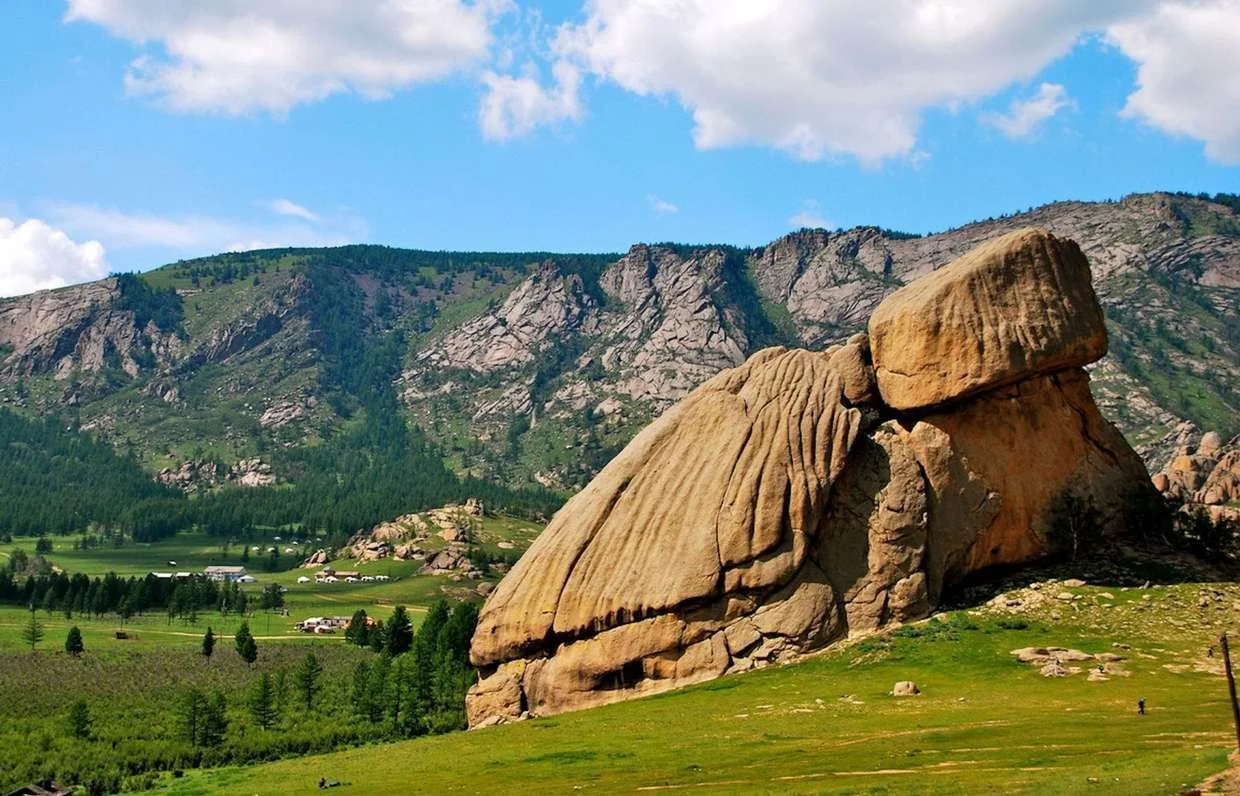 A large rock formation resembling a seated figure in a green valley with mountains and a partly cloudy sky in the background.