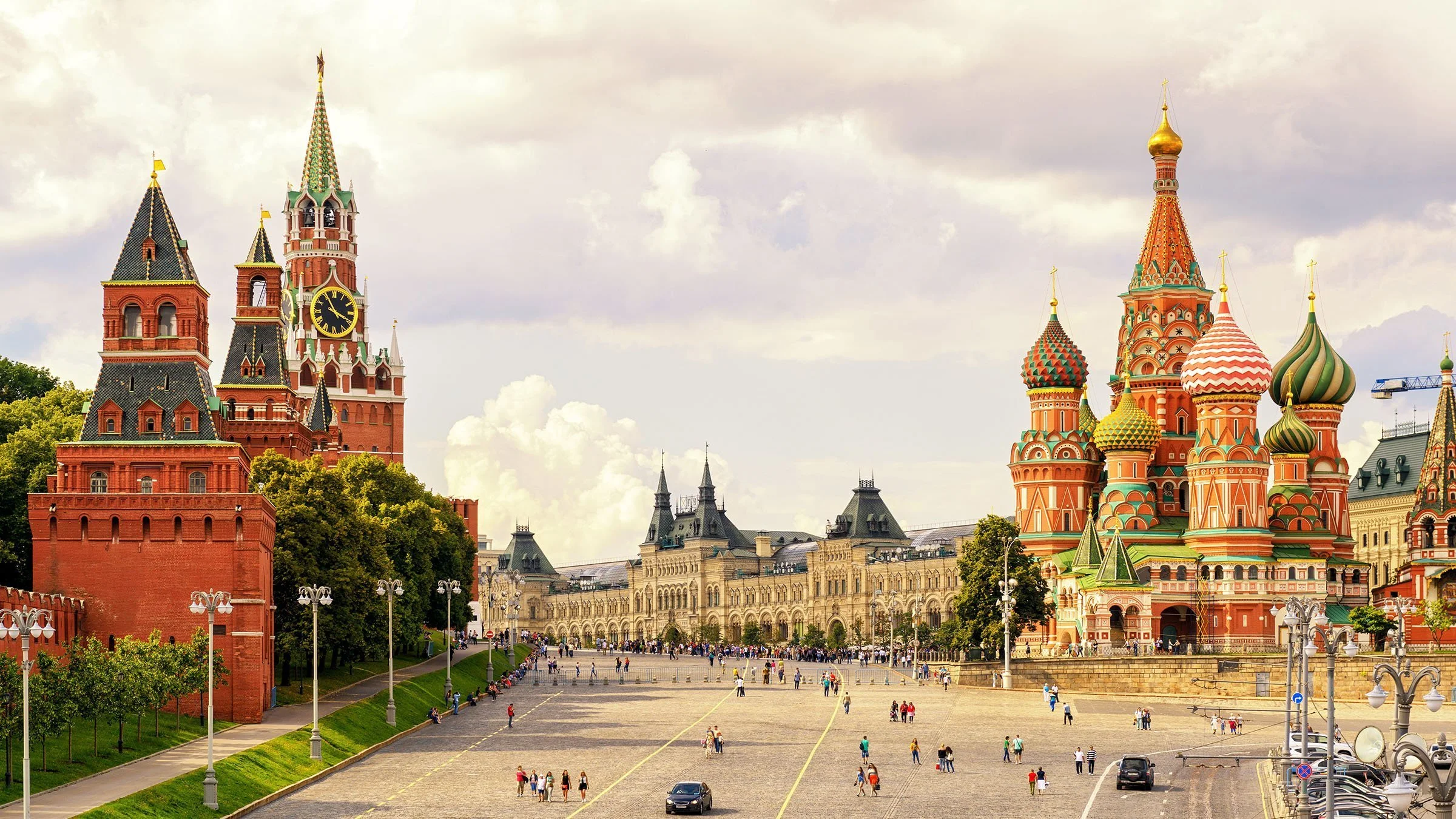 View of Red Square in Moscow, Russia, featuring the Spasskaya Tower with a clock on the Kremlin Wall on the left and St. Basil's Cathedral with colorful onion domes on the right, with people walking on the square.