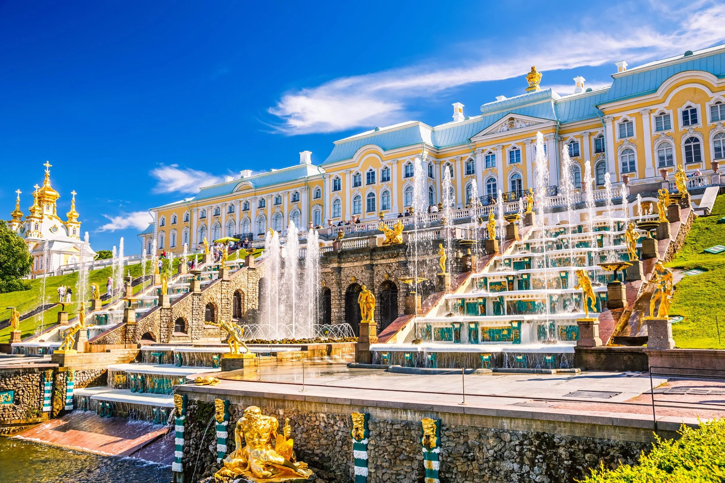 The Grand Palace in Екатеринбург, Russia, with a yellow and white baroque-style building, ornate water fountains with golden statues, and a bright blue sky.