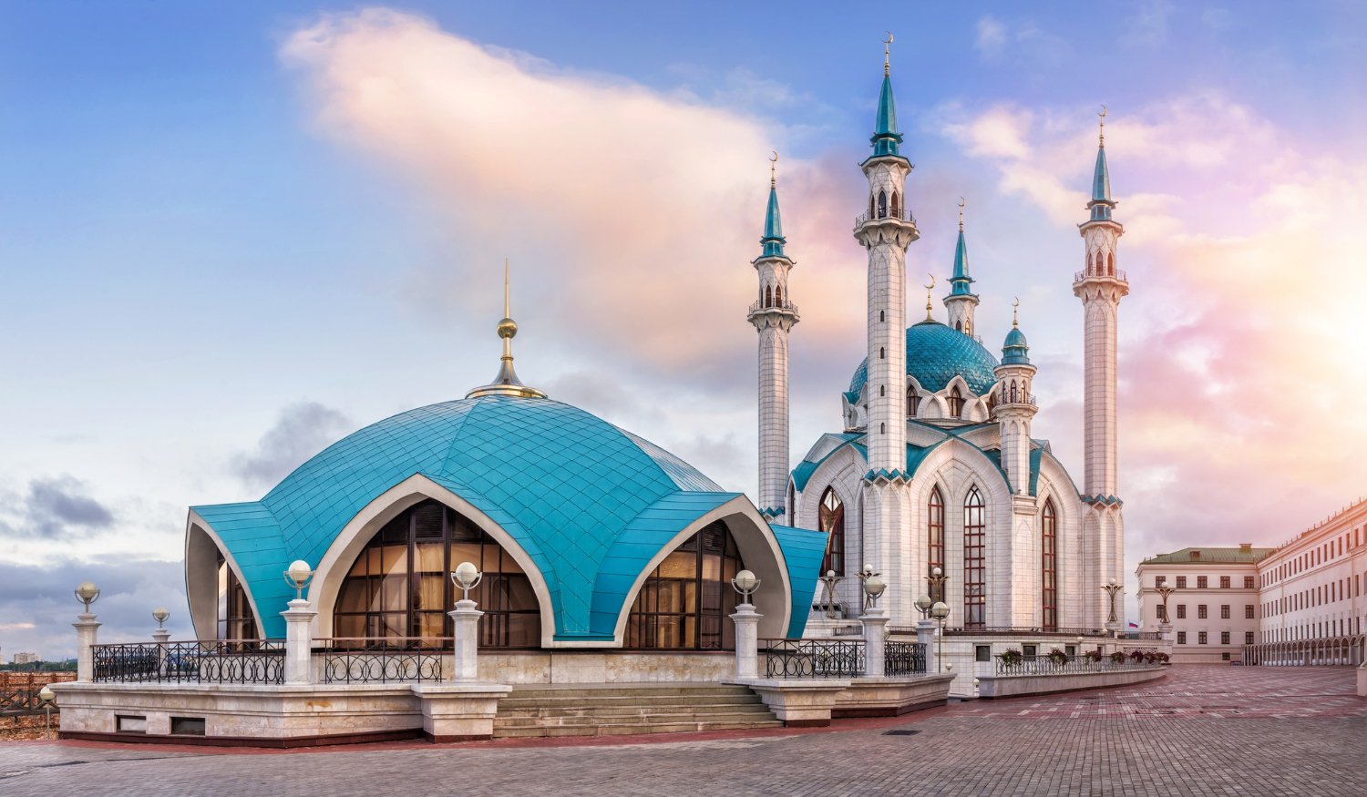 A mosque with a large blue dome and four minarets, set against a colorful sky with clouds during sunset.