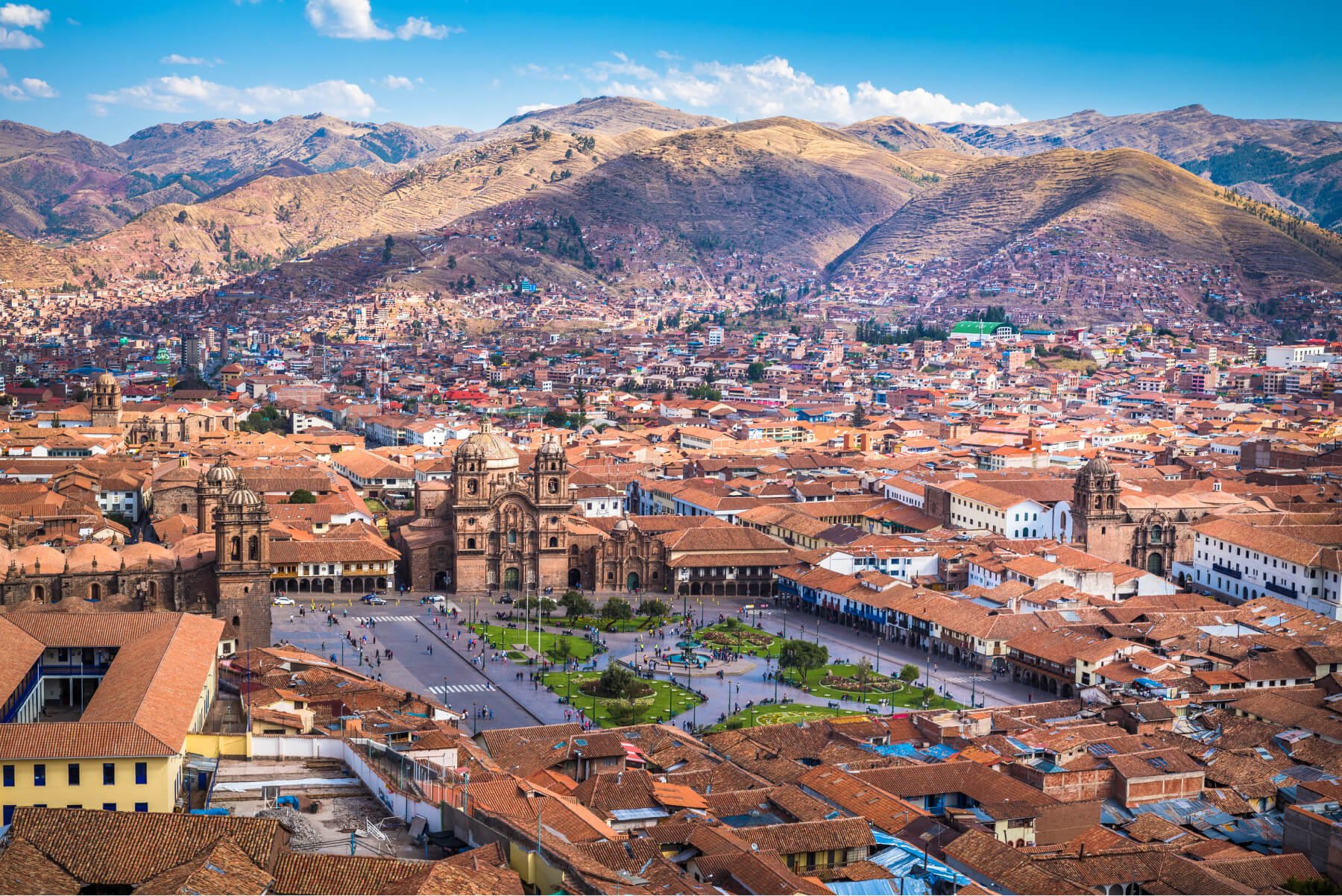 A panoramic view of a historic city with a large central cathedral, surrounded by red-tiled rooftops and a mountainous landscape in the background.