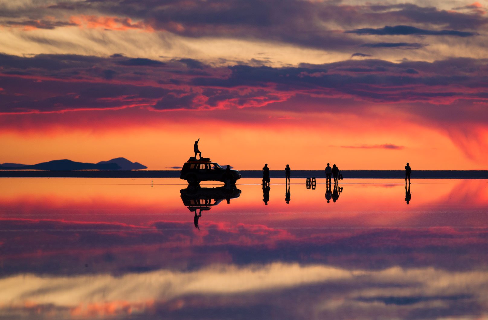 Silhouettes of people, a vehicle, and a person on top with a raised arm, set against a colorful sunset with reflections on water.