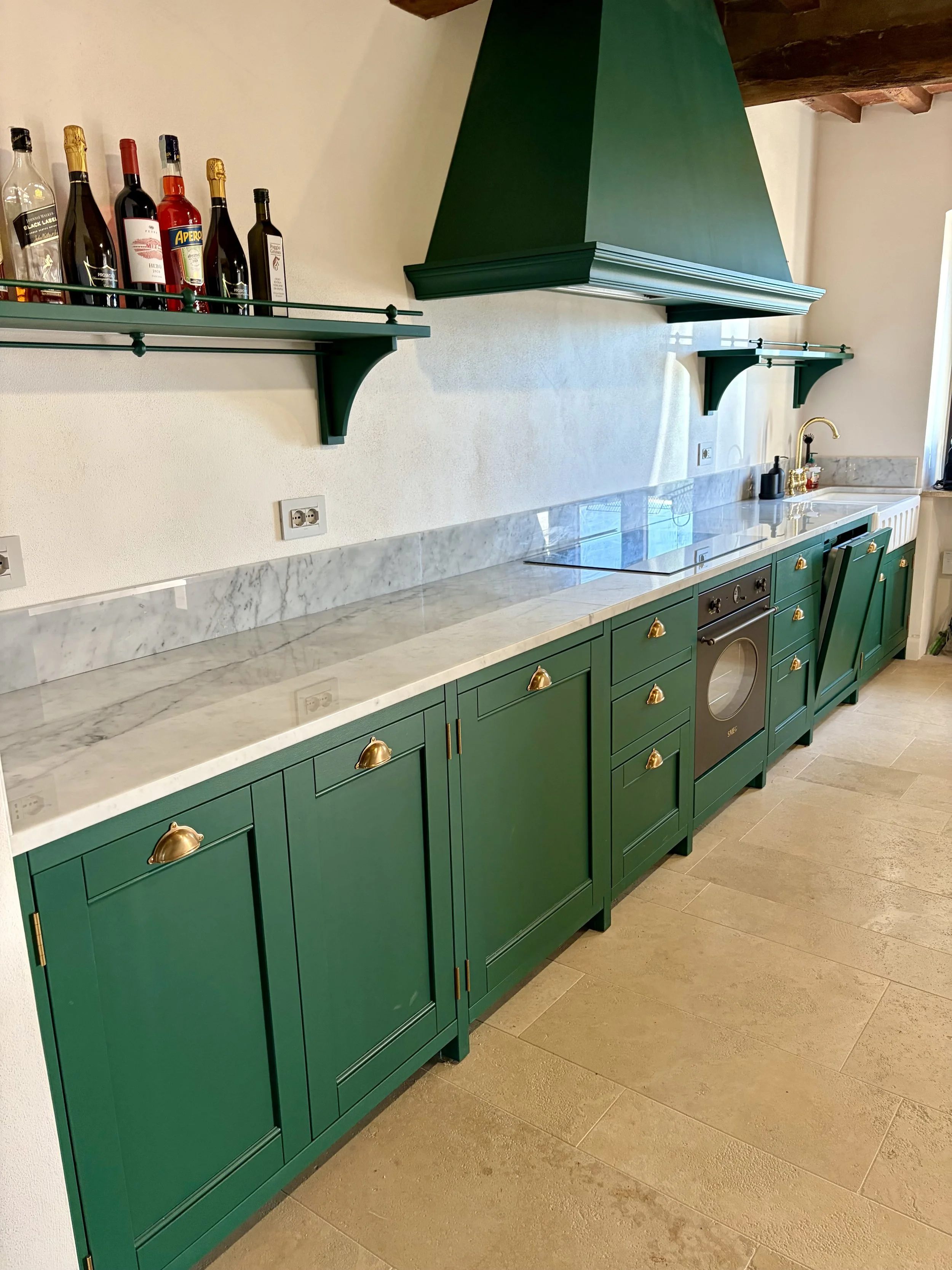 Green kitchen cabinets with gold handles, white marble countertop, black stovetop, and a sink with a gold faucet. Two green shelves with liquor bottles above the countertop. Beige tile flooring and a white wall with three electrical outlets.