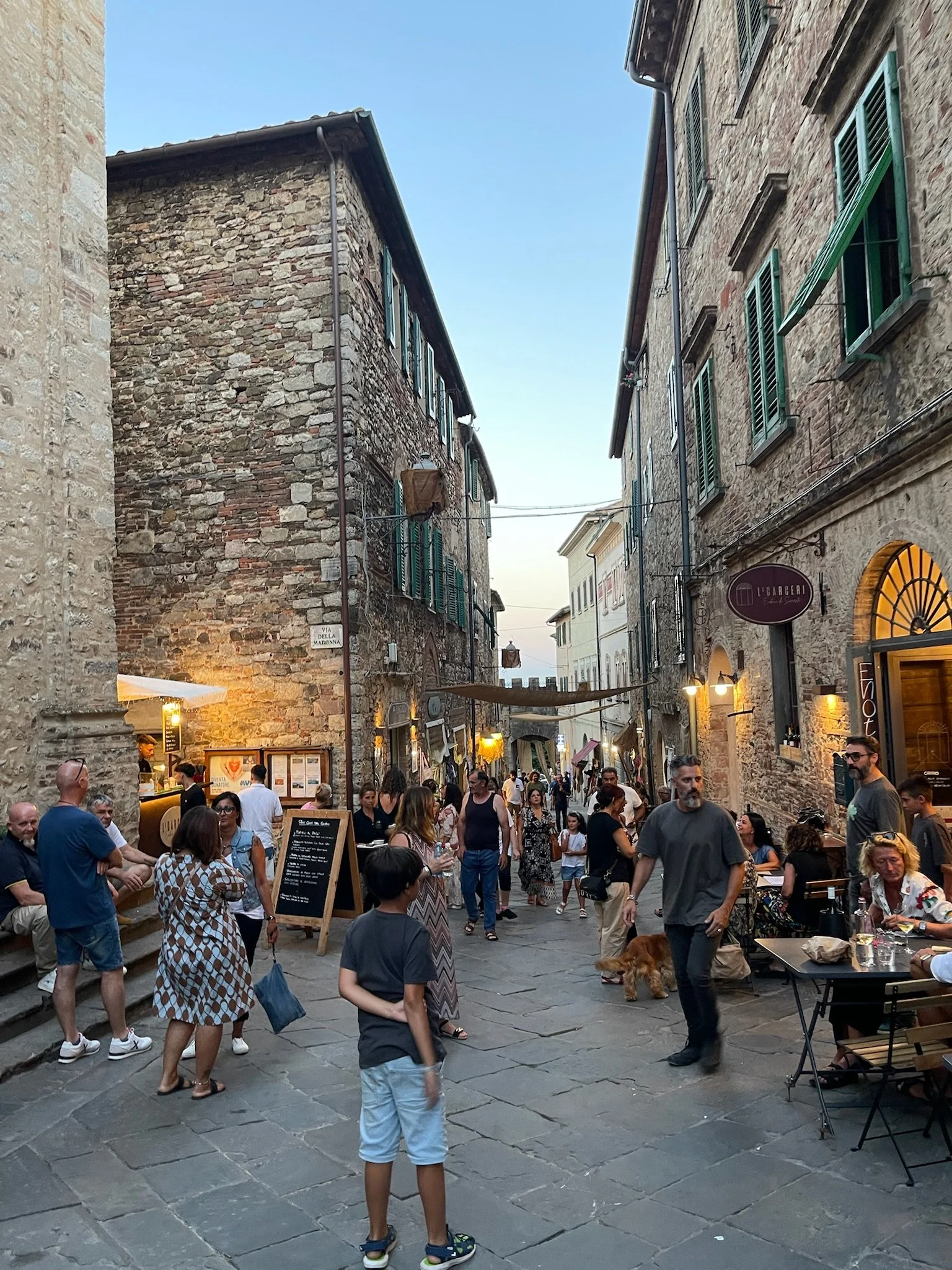 People walk and dine on a narrow cobblestone street in an historic European city, surrounded by stone buildings with green shutters and outdoor cafes.