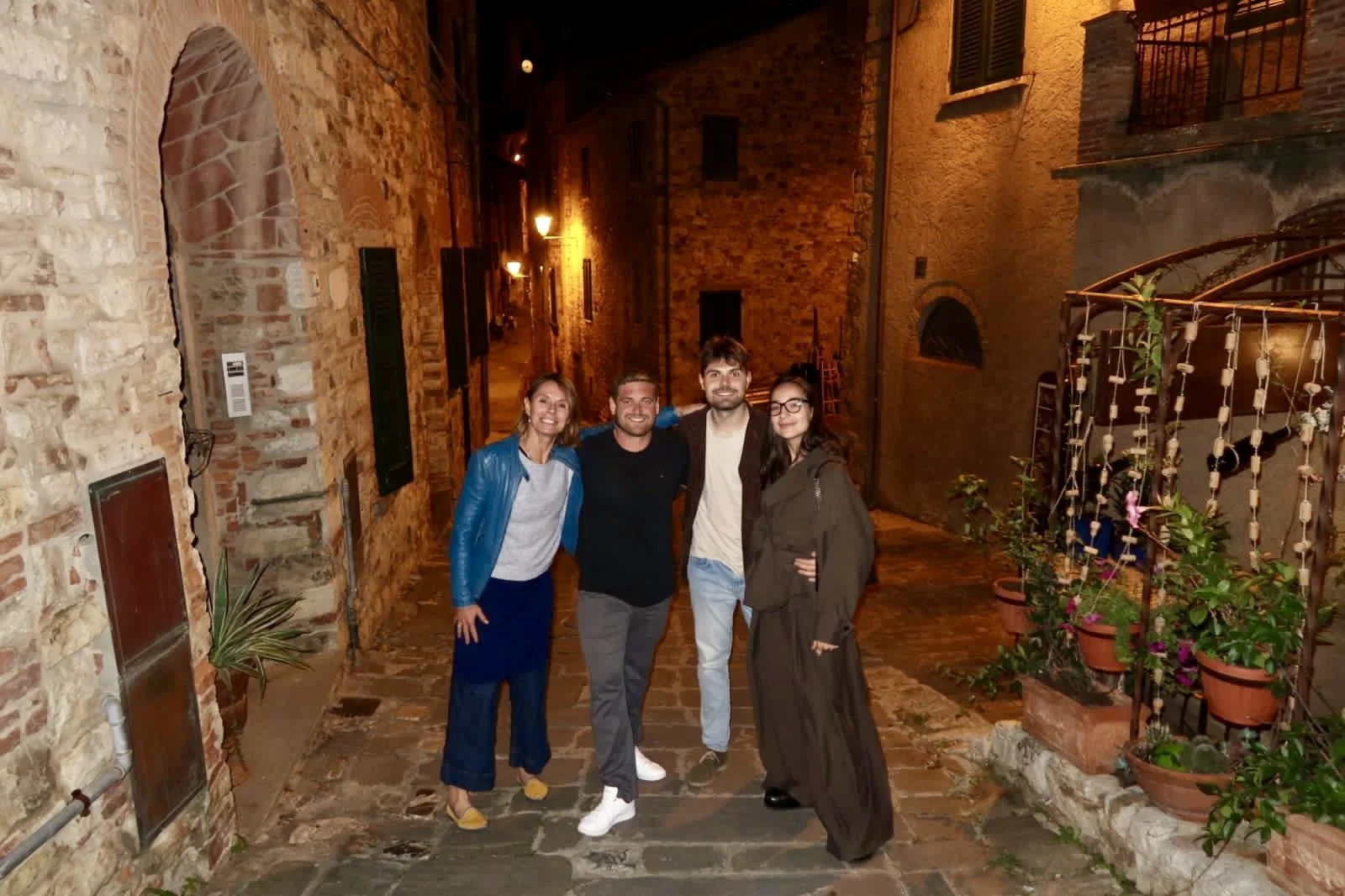 Four friends standing together in a narrow, cobblestone street at night, smiling, with brick buildings and potted plants around them.