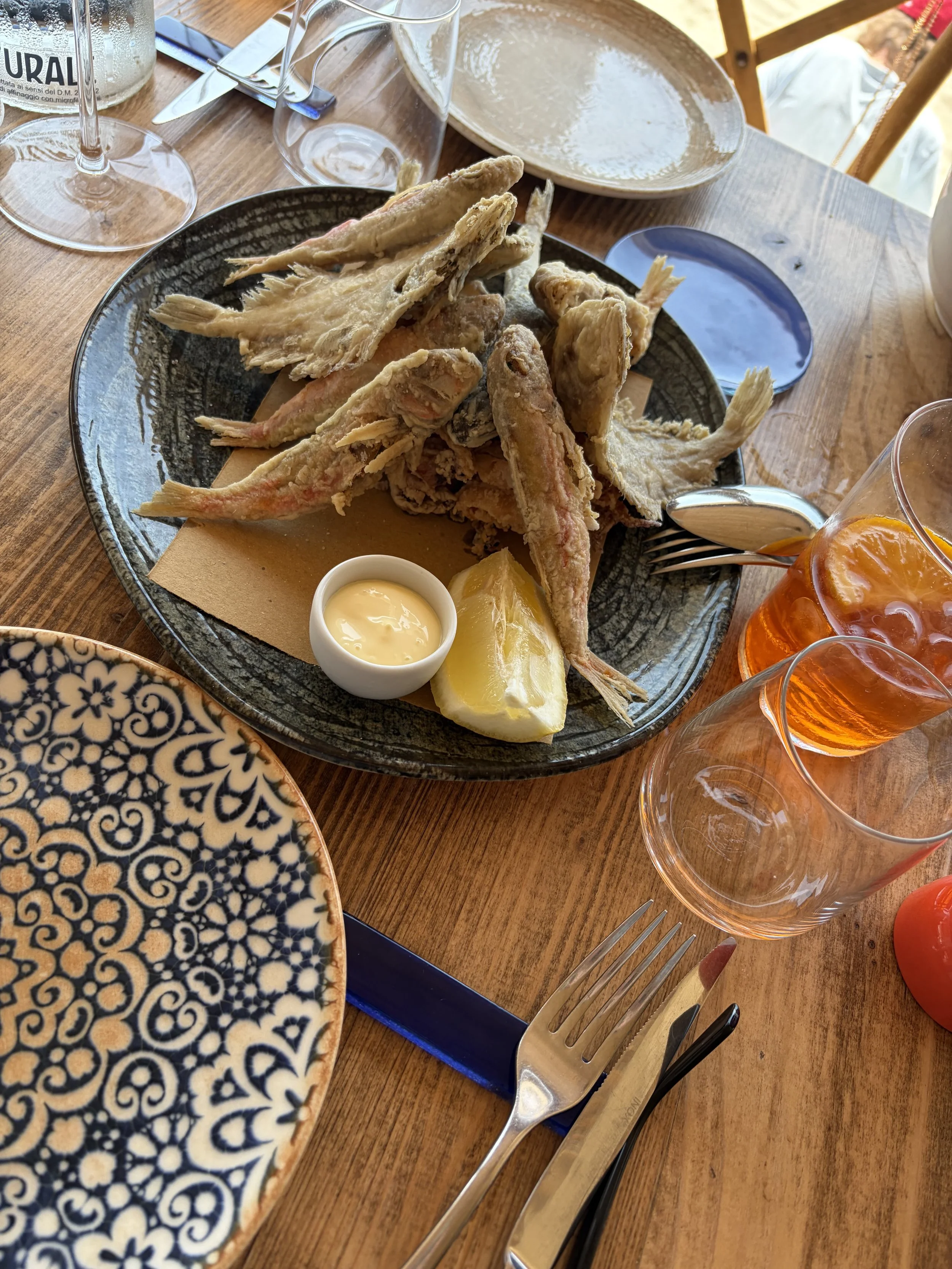 A plate of deep-fried fish with tartar sauce and lemon wedge, set on a wooden table with other glassware and dinnerware around.