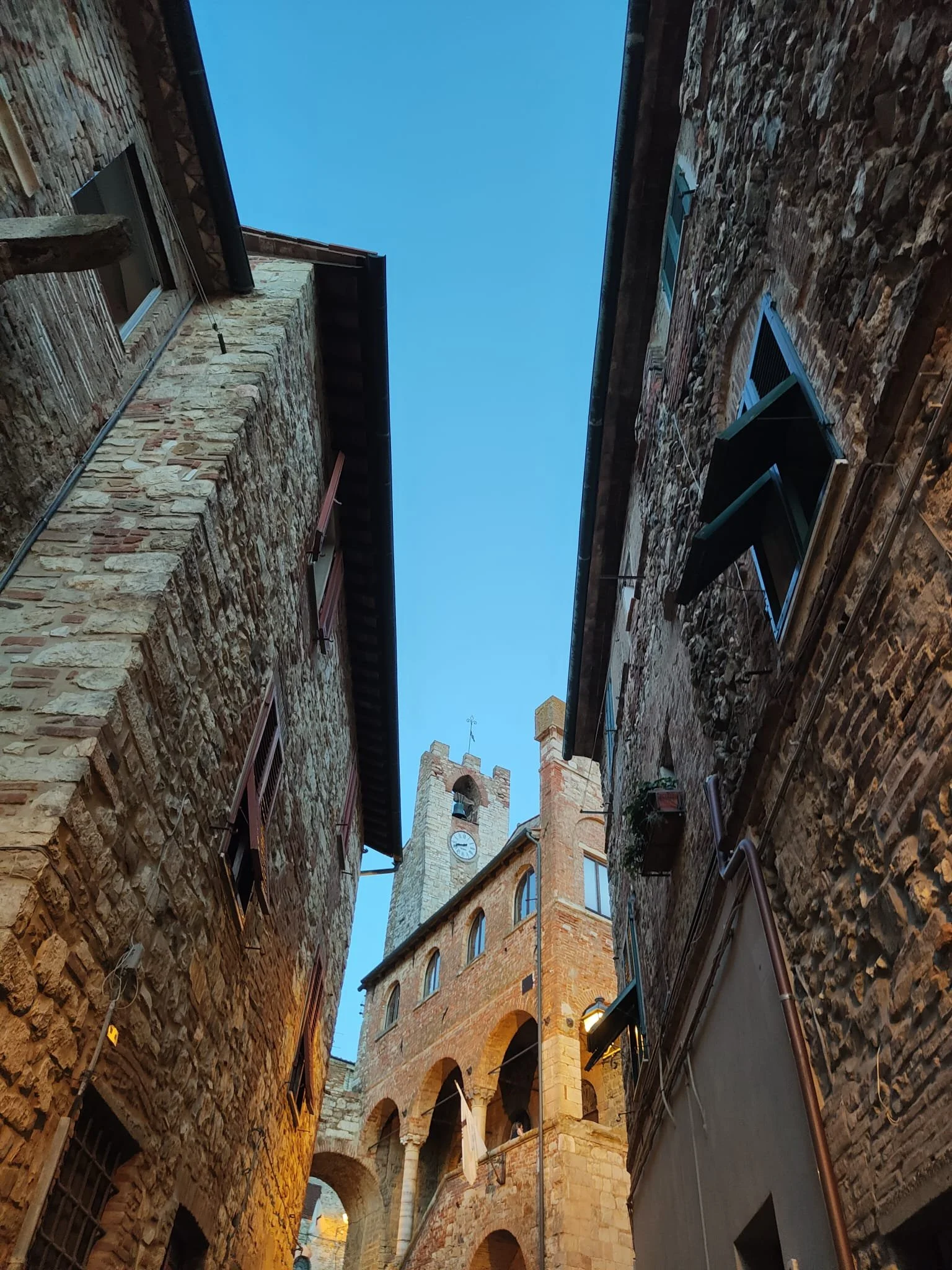 View looking up at the sky between two tall, old brick buildings with arched windows and a clock tower in the distance.
