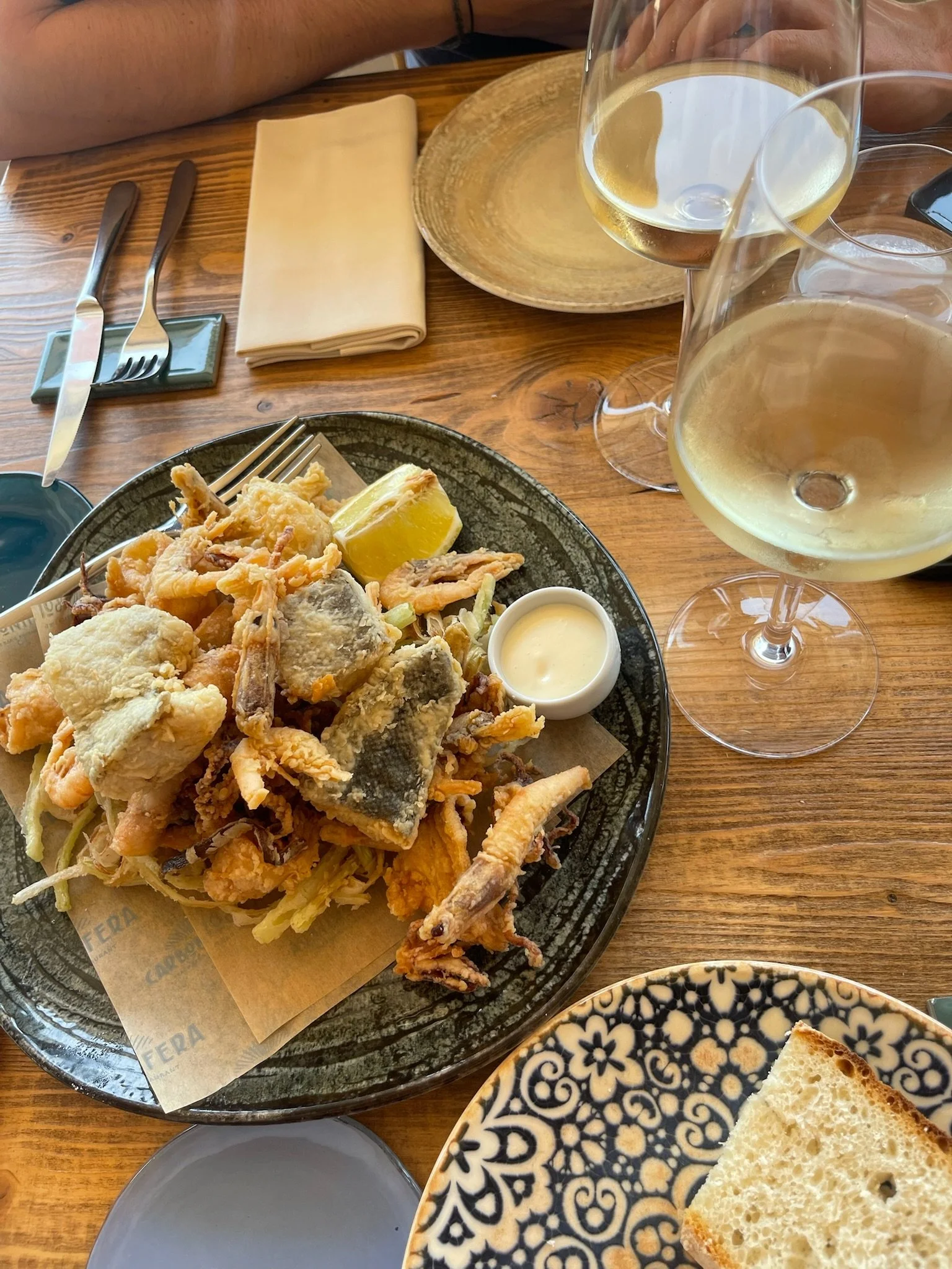 A plate of assorted fried seafood with lemon wedge and dipping sauce, two glasses of white wine, a napkin, and a plate of bread on a wooden table.